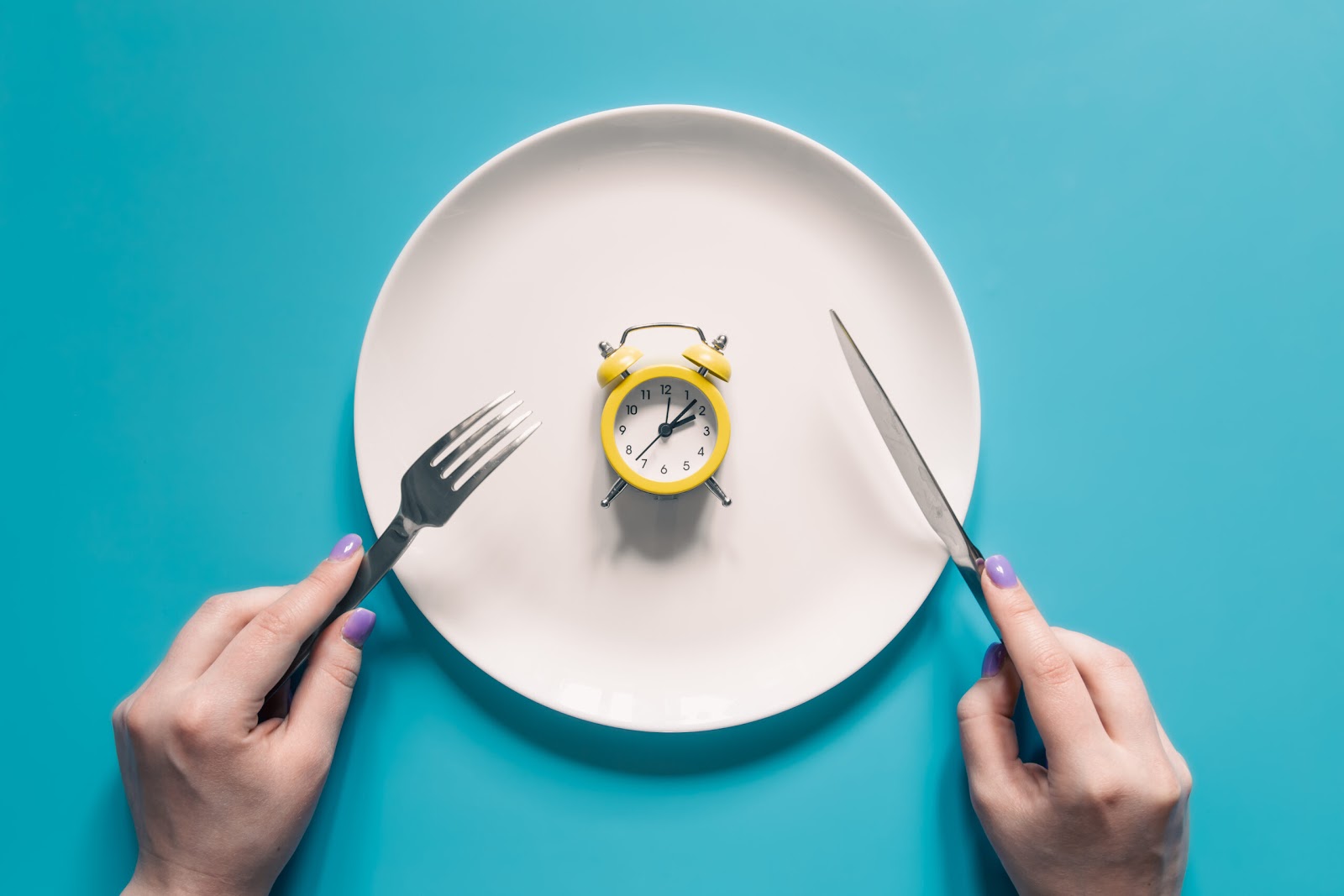 Hands holding knife and fork above alarm clock on a plate on blue background. Intermittent fasting, Ketogenic dieting, weight loss, meal plan, and healthy food concept.